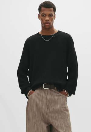 Man draagt een zwarte gestructureerde trui, beige wijde broek, zwarte riem, kettingarmbanden en een dunne ketting, staande met handen in de zakken.