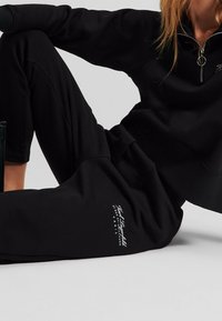 Person wearing black joggers and a matching black zip-up top, sitting on the floor with text logo visible on left thigh.