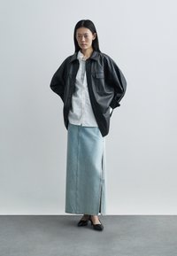 Woman standing with hands in pockets wearing black leather oversized jacket, white shirt, light blue denim maxi skirt, and black heels.