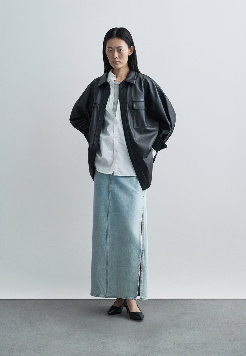 Woman standing with hands in pockets wearing black leather oversized jacket, white shirt, light blue denim maxi skirt, and black heels.