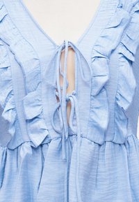 Φόρεμα από ανοιχτό μπλε ύφασμα με βολάν στη διακόσμηση και δύο δετές φιόγκους στο μπροστινό μέρος, με βαθύ V-neck και σούρες στη μέση.