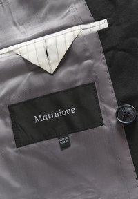 Melna auduma iekštelpa ar pelēku odere, kurai ir marķējums ar "Matinique" un izmēra detaļām. Pogas un salocīta balta kabata ar svītrām.