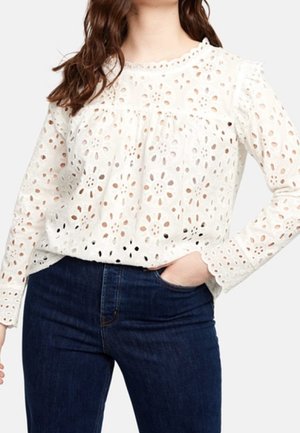 Biała bluzka z długim rękawem z ażurowej koronki z kwiatowymi wycięciami, noszona z ciemnoniebieskimi jeansami z wysokim stanem, jedna ręka w kieszeni, swobodna poza.