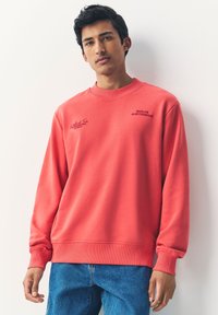 Felpa color corallo con girocollo e polsini a costine. Presenta la scritta grafica "Berlin Amsterdam" sul petto. Tessuto in misto cotone, vestibilità comoda.