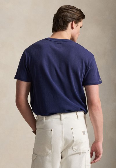 T-shirt blu navy a maniche corte con texture a coste, con scollo rotondo e piccolo logo sulla manica sinistra. Indossata con pantaloni color crema.