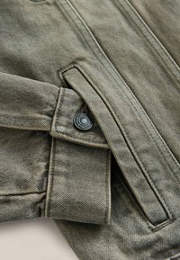 Zaļš džinsa jaka ar pogu aizdari, šuvumu detaļām un fakturētu auduma apdari. Dizainā ir apkakle un kabata.