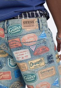 Jeansy z dżinsu z wieloma kolorowymi naszywkami i logo marki Guess Originals, noszone przez osobę o ciemnej karnacji i w koszuli w kratę włożonej do spodni.