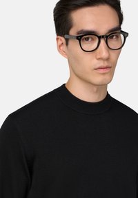 Pull en tricot noir avec un col rond haut, présentant une texture lisse et une coupe près du corps. Accentué par un mannequin portant des lunettes noires.