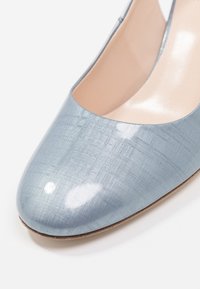 Vaaleansininen lakattu kengänpohja, jossa on teksturoitu pinta ja pyöreä kärki. Sisäosa on beige ja sileä, ja siinä on yksinkertainen muotoilu.