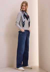 Graues Langarmshirt kombiniert mit weit geschnittenen dunkelblauen Jeans. Das Modell trägt einen gemusterten schwarzen Seidenschal und weiße Sneakers. Neutrale Hintergrund.