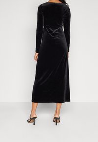 Robe en velours noire à manches longues avec une silhouette moulante, atteignant la longueur mi-mollet, dotée d'un design à dos dégagé et d'une texture lisse.