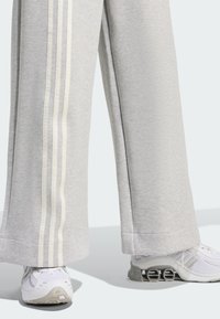 Pantalones deportivos de color gris claro con tres rayas verticales blancas en cada lado, con un corte relajado que se estrecha hacia el dobladillo. Combinados con zapatillas blancas.