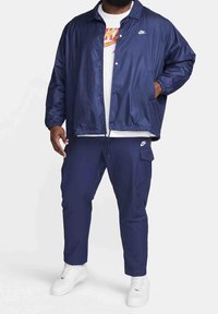 Mörkblå lättviktsjacka med tryckknappar, elastiska ärmslut och fickor, kombinerad med matchande byxor och vita sneakers. Vita logotypdetaljer.