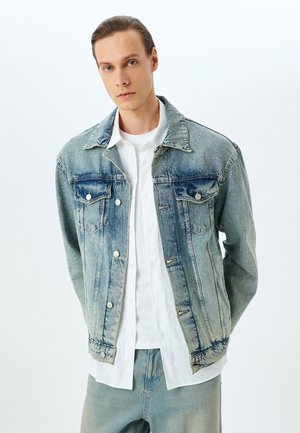 Hellblaue Jeansjacke mit einem Ausbleich-Finish, zwei Brusttaschen, Knopfverschluss und klassischem Kragen. Über einem weißen Hemd getragen.