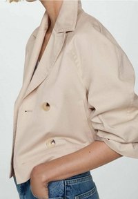 Veste courte beige en tissu lisse avec un design croisé à double boutonnage, présentant de gros boutons et des épaules structurées.