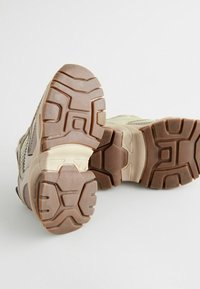 Beige atletiksko med teksturerede gummisåler, der har hævede mønstre og forstærkede hæle, designet til godt greb og holdbarhed.