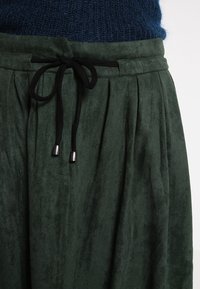 Falda de ante verde con cintura elástica ajustable, que presenta un lazo negro y detalles metálicos. La tela tiene una textura suave y lisa, con pliegues.