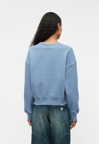 Ανοιχτό μπλε φούτερ με υφή, φαρδιά εφαρμογή, στρογγυλή λαιμόκοψη και μανίκια με χαμηλούς ώμους, συνδυασμένο με άνετα τζιν παντελόνια.