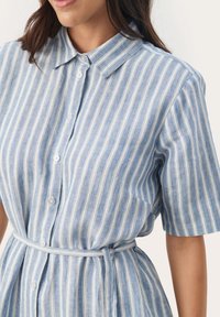 Robe chemise rayée bleu clair avec des manches courtes, fermeture à boutons sur le devant, col, et une ceinture en tissu à la taille. Fabriquée dans un matériau léger.