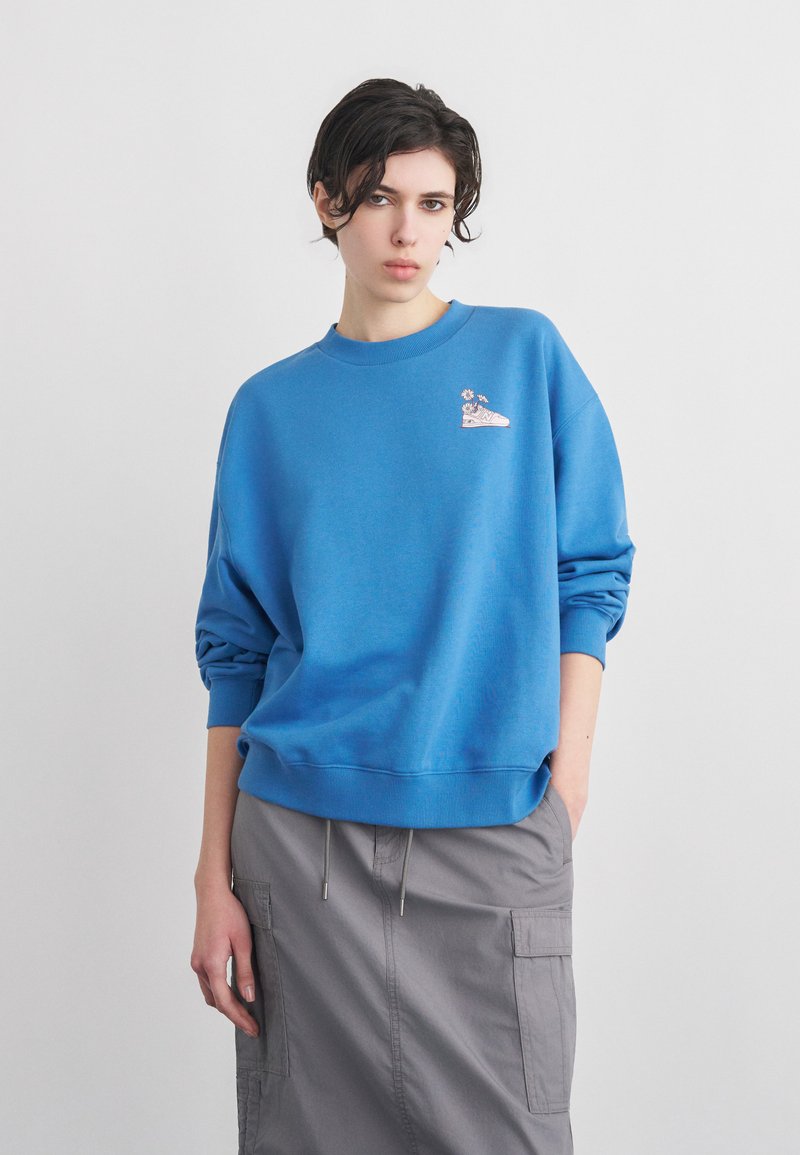 Νεαρό άτομο με κοντά σκούρα μαλλιά φοράει μπλε φούτερ με σχέδιο αθλητικού παπουτσιού και λουλουδιών, γκρι φούστα cargo, στέκεται μπροστά σε απλό φόντο.