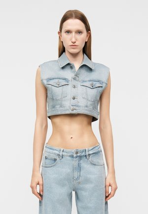 Frau trägt eine hellblaue ärmellose, kurz geschnittene Jeansjacke und passende hoch taillierte Jeans, steht vor einem schlichten grauen Hintergrund.