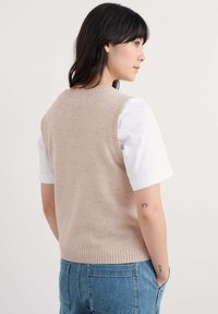 Beige kötött ujjatlan pulóver kerek nyakkivágással és bordás szegéllyel, fehér, rövid ujjú ing fölött viselve, kék farmernadrággal párosítva.