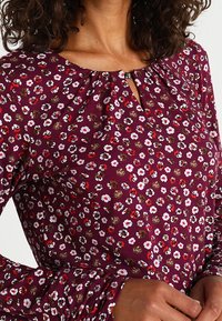 Blusa floreale viola con piccoli fiori bianchi e rossi, scollo rotondo con dettaglio a chiave e maniche lunghe con leggera arricciatura.