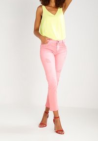 Top sin mangas de corte en v amarillo neón combinado con jeans rosa claro desgastados, con un corte corto y tacones rojos con tiras.