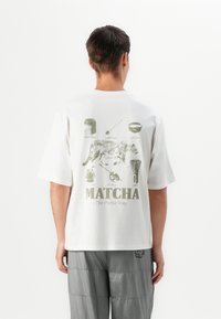 Bílý bavlněný tričko s zeleným grafickým potiskem kočky a různými předměty souvisejícími s matcha na zadní straně. Krátké rukávy, volný střih.