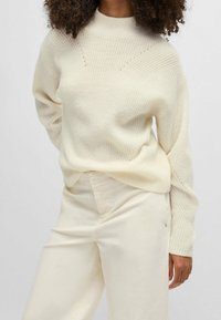 Femme portant un pull en maille côtelée crème et un pantalon large taille haute crème, se tenant avec une main détendue à ses côtés.
