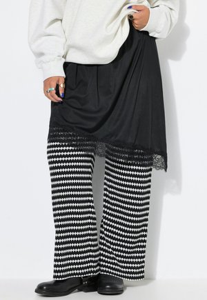 Person iført en hvid sweatshirt, sort nederdel med blonderkant, sort-hvide mønstrede bukser og sorte sko, med ringe på fingrene.