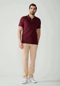 Bordeauxrotes Polohemd mit Reißverschlusskragen, kombiniert mit beigen Hosen und weißen Sneakers. Glatte Textur, enganliegendes Design, kurze Ärmel.
