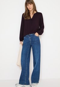 Ciemnofioletowa bluzka z długim rękawem, z dekoltem w kształcie V i guzikami z przodu, w połączeniu z szerokimi dżinsami w niebieskim kolorze. Modelka trzyma jasny szalik. Srebrne sneakersy.