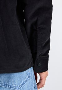 Camisa de pana negra con puños abotonados, que presenta una textura estriada y un dobladillo ligeramente curvado, combinada con denim azul claro.