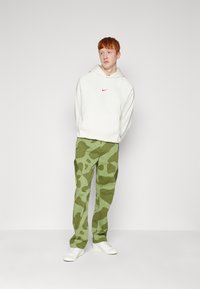 Balta bliuzono su raudonu Nike logotipu, žaliomis kamufliažo cargo kelnėmis ir baltomis kedais. Kasdienis stilius, medvilninė medžiaga, laisvas siluetas.
