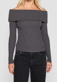 Grå off-shoulder topp med långa ärmar, ribbad textur, åtsittande form och kort längd, med en liten logotypetikett i kanten.