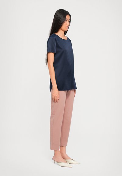 Woman with long dark hair wearing a navy short-sleeve top, light mauve cropped pants, and cream pointed-toe low heels standing sideways.