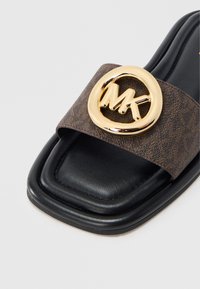 Sort flad sandal med firkantet tå, med en brun tekstureret rem dekoreret med en guld cirkulær logo. Glat læderfodbed.