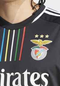 Czarna koszulka sportowa z okrągłym dekoltem, trzema białymi paskami na ramionach oraz kolorowym emblematem z orłem i gwiazdami.
