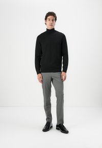 Schwarzer Rollkragenpullover, graue maßgeschneiderte Hose und glänzende schwarze Schuhe. Das Modell steht vor einem schlichten weißen Hintergrund. Klare Linien und moderne Passform.