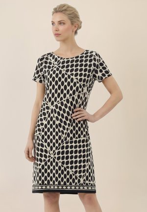 Blonde Frau steht mit einer Hand in der Hüfte, trägt ein knielanges schwarz-weißes Kleid mit großen ovalen Punkten und kurzen Ärmeln.