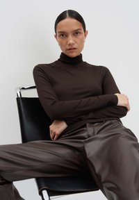 Bruine geribde coltrick met een paarse bruine leren wijde broek. Model zit op een zwarte stoel, met stijlvol haar en minimale accessoires.