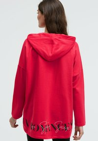 Rote Kapuzenjacke mit großer Kapuze, lässigem Schnitt und langen Ärmeln. Verfügt über schwarzen Text am unteren Rand und eine glatte Stoffstruktur.