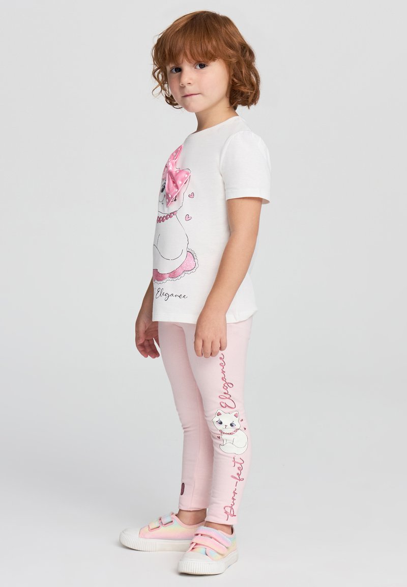 T-shirt bianco con un gatto cartoni animati con un fiocco rosa, abbinato a leggins rosa con un design di gatti. Calzature: sneakers dai colori pastello.