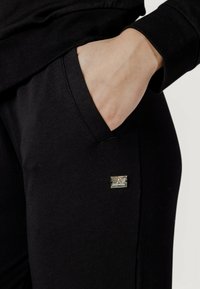 I pantaloni jogger neri in cotone presentano una tasca laterale e un piccolo tag in argento EA7 sul davanti, mostrando una texture liscia e un design aderente.