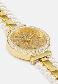 Gold wristwatch with pearl accents, round face, crystal-studded bezel, textured gold dial, and gold-tone hands. Brand name: Guess.