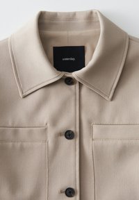 Beige jacket featuring a pointed collar, two buttoned chest pockets, and black buttons. The fabric has a smooth texture with neat stitching.
