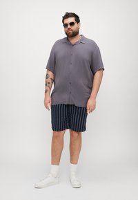 Camisa gris de manga corta con botones en el frente, combinada con pantalones cortos de rayas oscuras. El modelo lleva gafas de sol y zapatillas blancas, con tatuajes visibles.