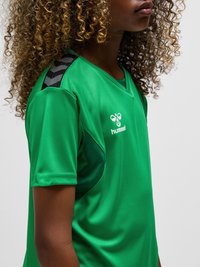 Młoda osoba z kręconymi włosami, ubrana w zieloną sportową koszulkę z czarnymi pasami w kształcie litery V na ramieniu i białym logo Hummel.