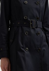 Marineblauwe trenchcoat met dubbele knoopsluiting, ceintuur op de taille en zijzakken. Gemaakt van een gladde stof met een satijnen afwerking.